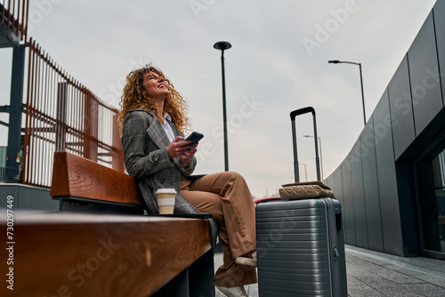 Sitting on a bench, a Caucasian woman uses her phone, holding a takeaway coffee with a suitcase beside her, patiently waiting for the bus.