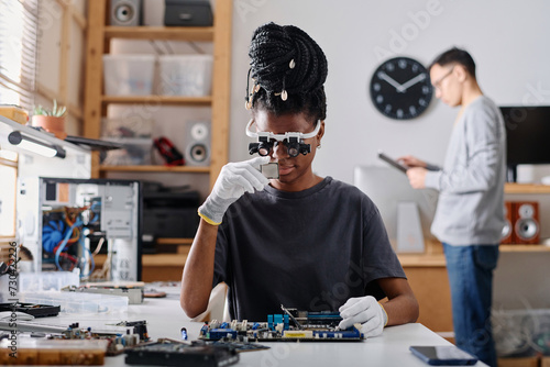 Female technician wearing glasses with loupes examining CPU, her male coworker standing on background