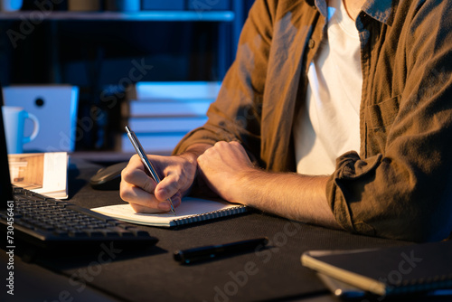 Cropped image of young businessman writing design thinking on paper for new project at neon light modern office. Thinking new next job sketching on notebook before sending present via email. Gusher.