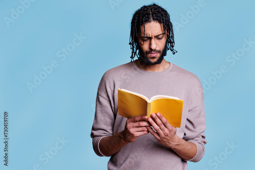Concentrated arab man reading paperback book with yellow cover. Focused young person with thoughtful expression studying literature, holding softcover textbook on blue background