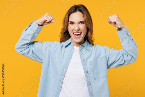 Young strong woman she wear blue shirt white t-shirt casual clothes showing biceps muscles on hand demonstrating strength power isolated on plain yellow background studio portrait. Lifestyle concept.