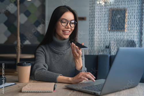 Happy business woman talking on speakerphone sitting at table with laptop, paper cup for coffee and notebook on it. Asian female sitting at office having telephonic conversation and smiling