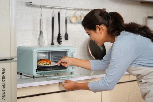 Indian happy smiling woman cooking burger by placing it on microwave oven at kitchen - concept of healthy eating, domestic lifestyle and technology