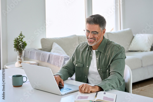 Smiling happy older mature middle aged professional man wearing eyeglasses looking at computer sitting at living room table, using laptop hybrid working, elearning, browsing online at casual home.