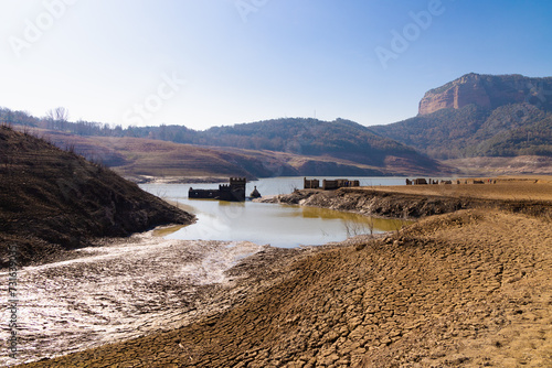 Un arroyo serpenteante fluye hacia el embalse de Sau, delineando las tierras cuarteadas y conectando con las estructuras sumergidas y los acantilados del Collsacabra en Osona, Cataluña.