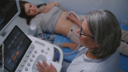 Female doctor conducts medical examination of stomach to female patient using sonography machine with digital monitor. Caucasian woman undergoes ultrasound diagnostics in modern clinic or hospital.
