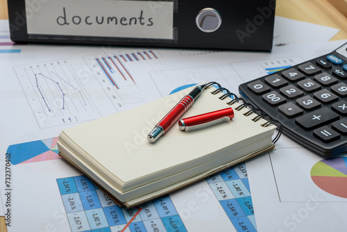 Segregator z dokumentami, kalkulator i papiery z danymi finansowymi, wykresami i analizami leżą na biurku