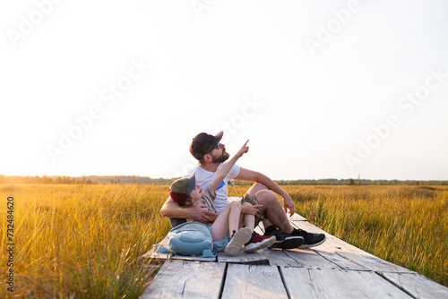 Father and son sitting on wooden boardwalk through swamp. Child looking and pointing his finger at the sky. Concept of ecotourism, hiking, vacation, travel with children. Selective focus.