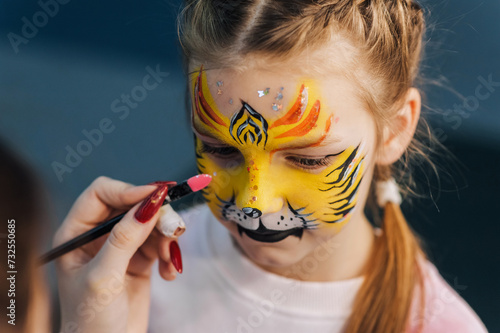 A professional artist draws with a hand with a brush, a colored pencil on the face of a child, a beautiful little girl, face painting, makeup, drawing at a party. Photography, creative process.
