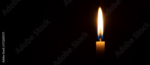 Single burning candle flame or light glowing on a small white candle on black or dark background on table in church for Christmas, funeral or memorial service with copy space.
