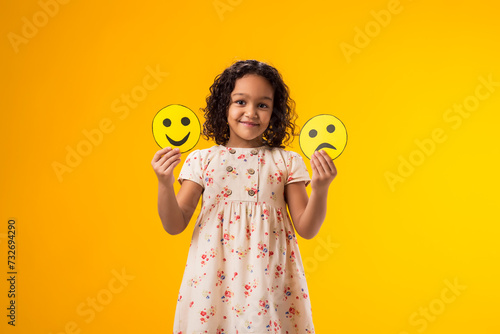 Smiling kid girl holding sad and happy emoticons in hands. Mental health, psychology and children's emotions concept