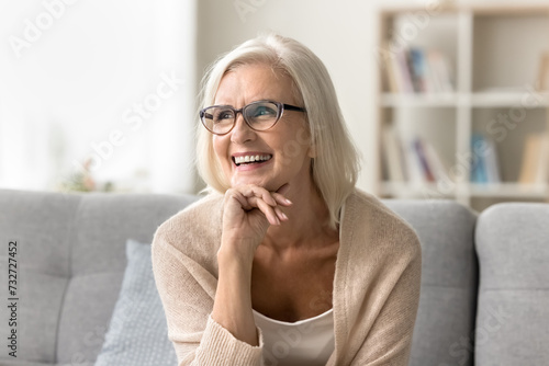 Happy blonde old senior woman in elegant glasses looking away home portrait. Cheerful positive elder female model leaning chin on hand, thinking on retirement with toothy smile