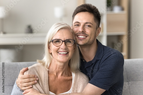 Cheerful caring adult son hugging happy blonde senior mother with love, support, looking at camera with perfect toothy smile, laughing. Elderly mother and grown male child headshot portrait