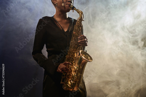 Cropped shot of elegant Black young woman playing saxophone in jazz music club with smoke effects copy space