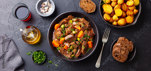 Beef meat and vegetables stew in black bowl with roasted baby potatoes. Dark background. Top view.