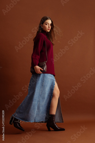 Fashionable confident woman wearing burgundy color knitted sweater, blue denim maxi skirt, leather pointed toe ankle cowboy boots, posing on brown background. Full-length studio fashion portrait