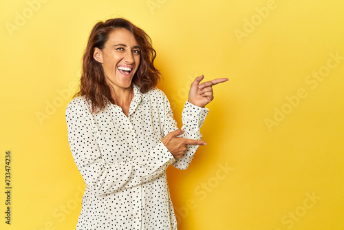 Middle-aged woman on a yellow backdrop pointing with forefingers to a copy space, expressing excitement and desire.
