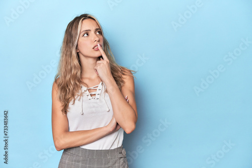 Blonde young caucasian woman in blue studio looking sideways with doubtful and skeptical expression.