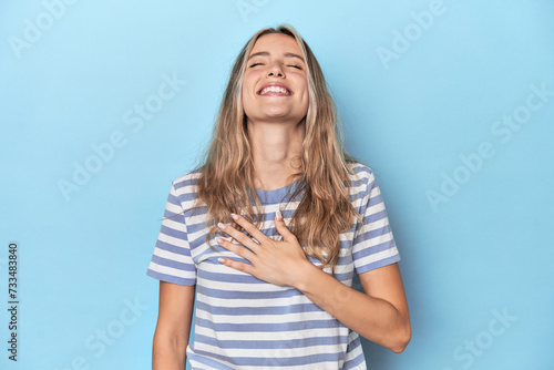 Blonde young caucasian woman in blue studio laughs out loudly keeping hand on chest.