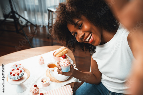 Beautiful young African blogger presenting piece of cupcake in concept special cuisine with selfie on smartphone. Content creating of social media with favorite sweets bakery dish. Tastemaker.