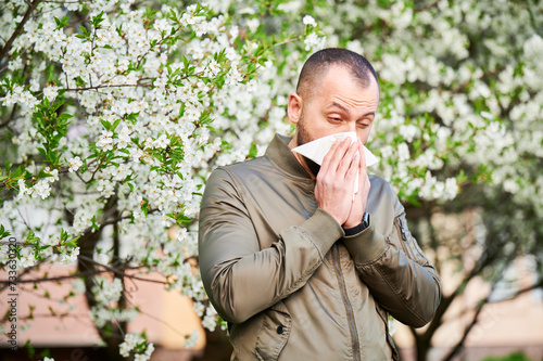 Man allergic suffering from seasonal allergy at spring in blossoming garden at springtime. Man sneezing and blowing nose using nasal handkerchief in front of blooming tree. Spring allergy concept.