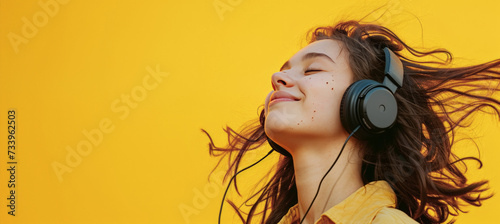 Bella mujer joven de 20 años sonriente de pelo largo suelto, escuchando música con unos auricularess negros y camisa amarrilla, sobre fondo amarillo con espacio vacio para publicidad