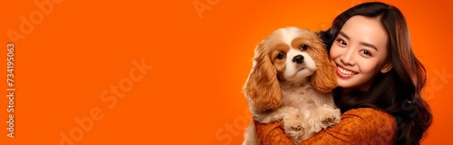 The image features a cheerful young woman with long hair, dressed in a warm orange sweater, tenderly holding a fluffy, beige and white Cavalier King Charles Spaniel. Both are set against a vibrant ora