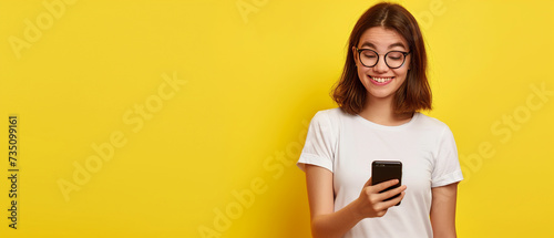 Mulher jovem surpresa em camiseta branca, segurando o smartphone e olhando para a câmera isolada em fundo amarelo