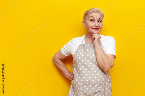 thoughtful old grandmother housekeeper in an apron plans and thinks on a yellow isolated background, elderly woman housewife imagines and dreams looks away