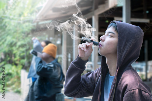 Asian male teenager is smoking electronic cigarette or vapor and drinking alcohol with friends in secret area, soft focus, daily life problems and bad habit and behavior of teenagers concept.