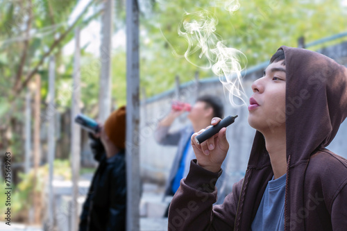 Asian male teenager is smoking electronic cigarette or vapor and drinking alcohol with friends in secret area, soft focus, daily life problems and bad habit and behavior of teenagers concept.