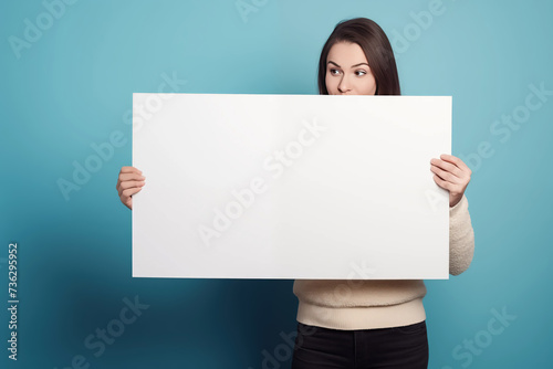 A girl or woman standing in a studio holding a blank sign for your own message of protest, sale, promotion or other against a colour, coloured, colorama backdrop green blue