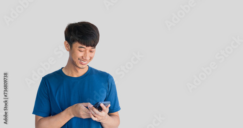 A young Asian man in his 20s wearing a blue t-shirt is happily playing on a mobile phone isolated on a white background. Typing and chatting with friends via smartphone. Online social concept.