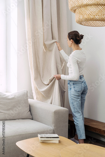 Woman standing in living room fixing linen curtains on window