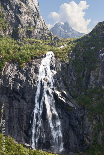 Valefossen waterfall flows vigorously down the cliffs of Lofoten Islands, Norway, amidst lush foliage and under a dynamic summer sky.