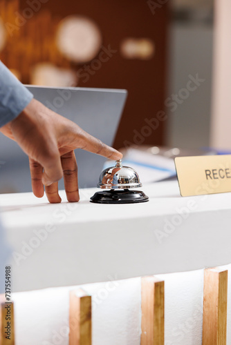 Close up of male hand pressing down front desk bell button while standing at empty reception counter, selective focus. Guest arrived at hotel, waiting for receptionist to check-in