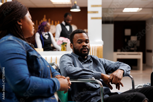 Young African American man in wheelchair in hotel lobby with able-bodied girlfriend, waiting for accessible room, handicapped guy traveling with mobility impairment. Accommodations for disabled