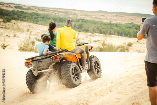 Tourist taking picture of family riding ATV all-terrain vehicle with angled shoulder tread tire improved grip, traction, front shock absorber suspension quad bike, white sand dunes, Mui Ne