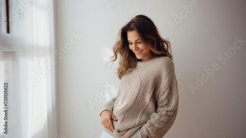 A smiling pregnant woman wearing a cozy knit sweater, cradling her belly with love and anticipation, against a minimalist white wall.