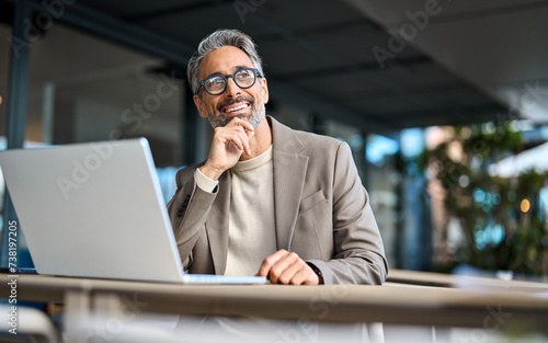 Happy mature business man entrepreneur using laptop computer outside office looking away. Smiling busy middle aged professional male entrepreneur sitting at table outdoors hybrid working. Copy space