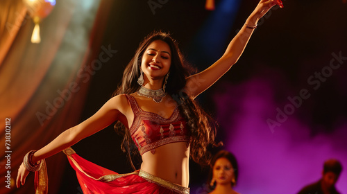 A South Asian woman with long brown hair, in a vibrant red sari, joyfully dances on stage