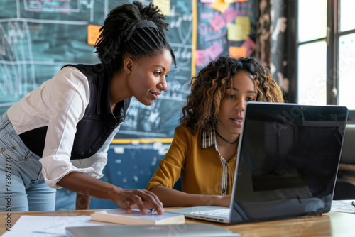 I have question. Black woman teacher stand by desk of mixed race female student look on pc screen help to solve problem. Skilled biracial team leader check online work result of young lady apprentice