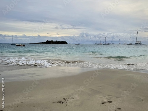 captivating scene in Guadeloupe with a sandy beach bordered by a distant islet and sailboats on the horizon. Waves dance under overcast skies, creating a mysterious ambiance in this tropical enclave 