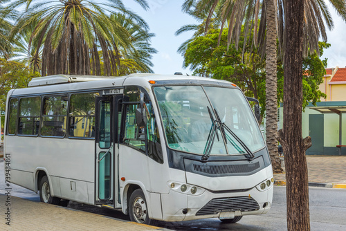 Close-up view of white passenger minibus at the Willemstad, Curacao bus stop, with background of tropical trees and blue sky.