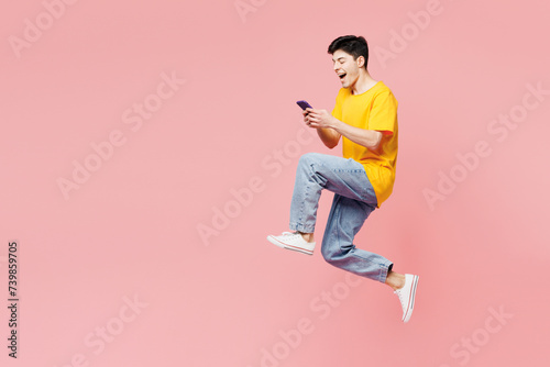Full body side profile view excited young man wear yellow t-shirt casual clothes jump high hold use mobile cell phone isolated on plain pastel light pink background studio portrait. Lifestyle concept