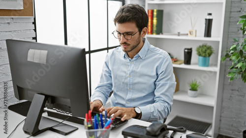 A focused hispanic man with a beard works at a computer in a modern office setting.