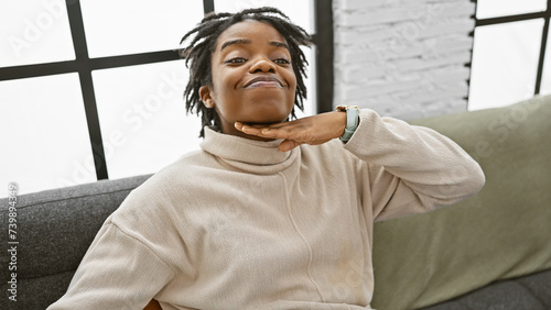 Portrait of a young woman with dreadlocks making a cutthroat gesture indoors on a sofa.