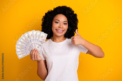 Portrait of satisfied cheerful girl with perming coiffure holding money showing thumb up get salary isolated on yellow color background