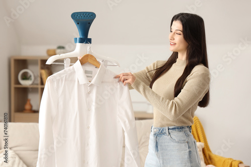 Pretty young woman with modern garment steamer and shirt at home