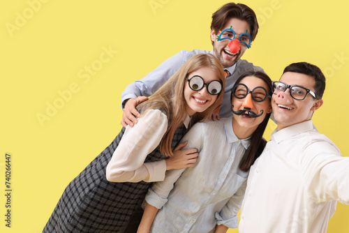 Business colleagues in funny disguise taking selfie on yellow background. April Fools' Day celebration